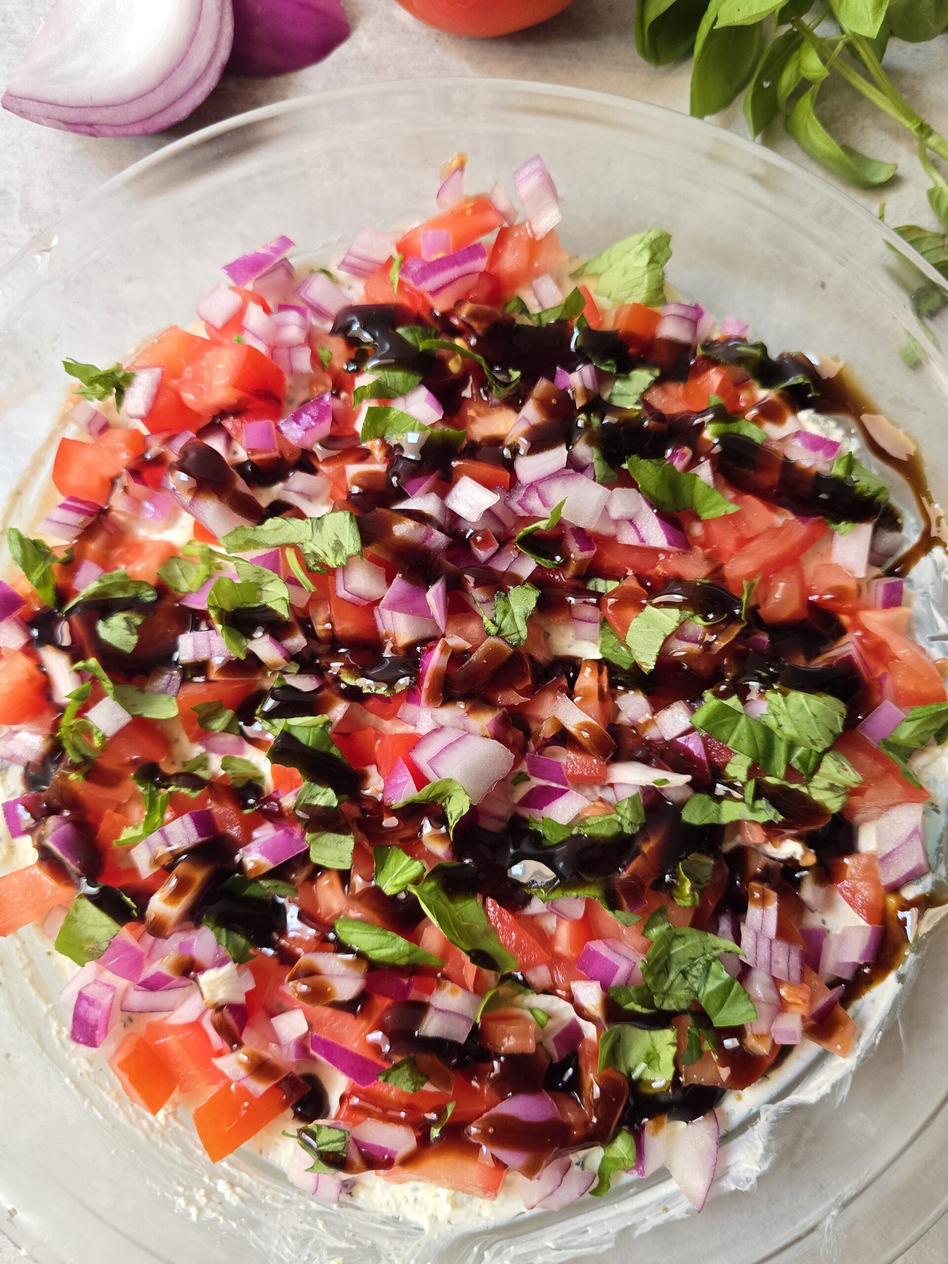 Creamy gluten free Caprese dip topped with diced tomatoes, red onion, basil, and balsamic glaze in a clear pie dish.