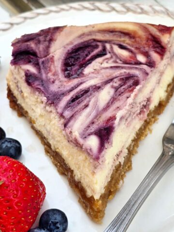 Slice of gluten free berry swirl cheesecake with creamy filling, strawberry and blueberry swirls, and a gluten free graham cracker crust.