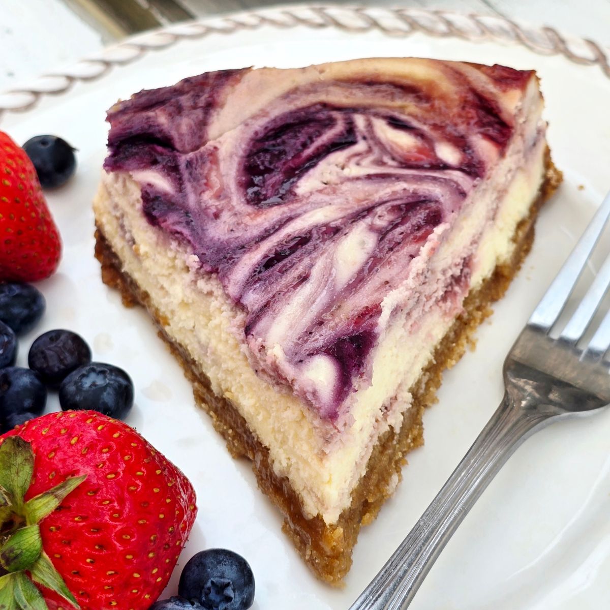 Slice of gluten free berry swirl cheesecake with creamy filling, strawberry and blueberry swirls, and a gluten free graham cracker crust.