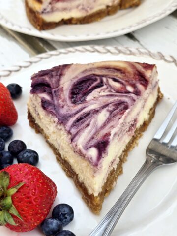 Slice of gluten free berry swirl cheesecake with creamy filling, strawberry and blueberry swirls, and a gluten free graham cracker crust.